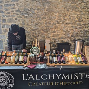 Stands de L'Alchymiste : bouteilles de liqueurs alignées, vendeur penché, mur de pierre.