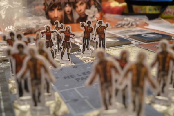 Des figurines de personnages de zombies en carton se trouvent au premier plan, disposées sur un plateau de jeu. Les zombies sont représentés dans des poses menaçantes, certains levant les bras.