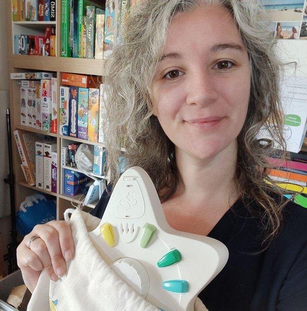 Eve Oika Oika. Une femme souriante avec des cheveux gris tenus lâchés tient un appareil électronique dans une main et une sacoche en tissu dans l'autre. En arrière-plan, on aperçoit une étagère remplie de jeux de société et de divers objets colorés.