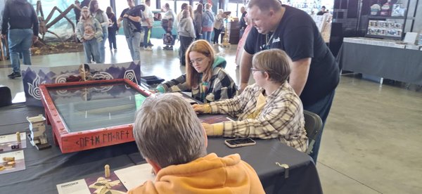 Des personnes jouent à un jeu de société sur une table lors d'un événement. On voit un grand plateau de jeu décoré, entouré de plusieurs joueurs, dont une jeune fille aux cheveux roux et deux autres personnes, concentrées sur le jeu.