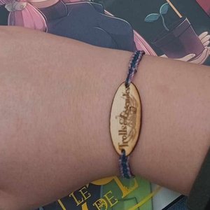 Un bracelet en bois gravé avec le texte "Trolls & Légendes" est porté au poignet d'une main. En arrière-plan, on aperçoit une couverture de livre illustrée avec une femme tenant une plante. L'image évoque l'univers du festival en juillet 2025.