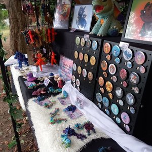 Stands de badges, peluches et bijoux fantaisie colorés sous une tente, décor féerique.