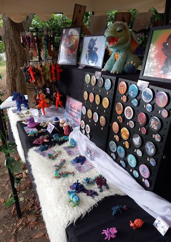 Stands de badges, peluches et bijoux fantaisie colorés sous une tente, décor féerique.