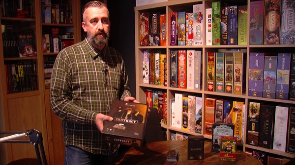 Vincent Joassin. Un homme avec une barbe, portant une chemise à carreaux, se tient devant une étagère remplie de jeux de société. Il tient dans une main une boîte de jeu intitulée "Contract".