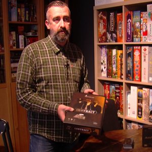 Vincent Joassin. Un homme avec une barbe, portant une chemise à carreaux, se tient devant une étagère remplie de jeux de société. Il tient dans une main une boîte de jeu intitulée "Contract".