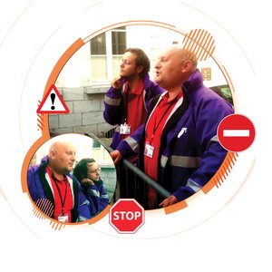 Deux hommes, vêtus de vestes violettes et de t-shirts rouges, se tiennent près d'une barrière. L'un d'eux, chauve et parlant, paraît concentré, tandis que l'autre, avec des lunettes et un regard pensif, regarde vers le haut. En arrière-plan, on aperçoit u