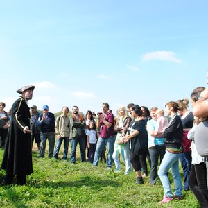Un homme en costume historique, avec un chapeau, se tient debout devant un groupe de personnes rassemblées, écoutant attentivement. Les participants, d’âges divers, se tiennent sur un chemin herbeux sous un ciel bleu. Certaines personnes semblent interagi