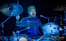 Vibe intense: un batteur des JOLLY JACKERS, bandana verte et veste bleu sombre, frappe furieusement la batterie sur scène. Cymbales et futs brillants sous les feux bleus, micro à portée. Ambiance live énergique du Festival 2026, hub Concerts.