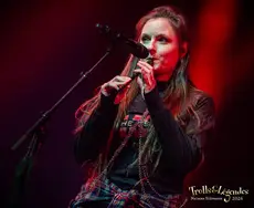 Chanteuse des JOLLY JACKERS sur scène, micro à la bouche, vêtue d’un pull sombre et d’un jean à carreaux noué autour des hanches. Lumières rouges intenses, ambiance live au hub Concerts du Festival 2026.