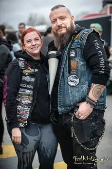 Ambiance Générale: Festival 2026. Deux passionnés posent ensemble: femme en hoodie noir décoré de patches multicolores et leggings graphiques, homme barbu en gilet jean chargé de patches et de chaînes, tous deux souriants devant la scène.