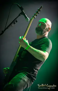 SAOR en plein live sur scène, guitare électrique en avant, micro prêt à vibrer. Homme barbu et rasé, t-shirt noir, éclairage vert enveloppant. Ambiance dense du Festival 2026 au hub Concerts.