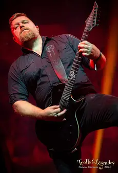 Guitariste posé en burg avec chemise noire, barbe marquée, en plein solo sur scène. Guitare noire à manche moderne et accessoire argenté. Lumières rouges enveloppantes, ambiance rock électrique; scène du Festival 2026, hub Concerts.