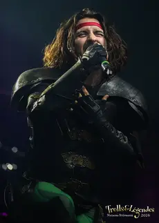 GLORYHAMMER en plein dans le feu des projecteurs: silhouette héroïque en armure sombre, harnais cuir, cape billowing. Il crie dans le micro, headband rouge, énergie furieusement théâtrale sur scène lors d’un Festival 2026. Concerts, ambiance épique.