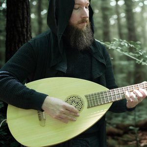 Chansons et Dragons. Un homme avec une longue barbe joue d'un luth dans une forêt verdoyante. Il porte une cape noire avec un capuchon, et la lumière filtre doucement à travers les arbres, créant une ambiance mystique.