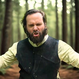 Chansons et Dragons. Un homme avec une barbe, portant une chemise jaune et un gilet noir, s'exprime avec intensité au milieu d'une forêt. Il a les bras écartés et semble passionné. Des arbres majestueux forment un paysage verdoyant en arrière-plan.