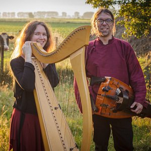 Fyndus. Deux musiciens posent dans un champ verdoyant baigné par la lumière dorée du coucher de soleil. La femme, souriante, tient une harpe, tandis que l'homme porte un instrument à cordes. En arrière-plan, on aperçoit des vaches au pâturage.