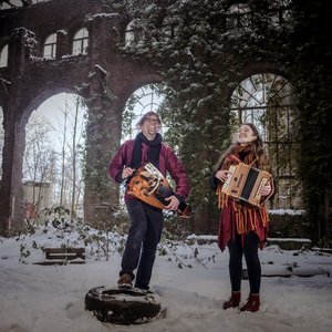 Fyndus. Deux musiciens se tiennent dans un ancien bâtiment en ruine recouvert de neige. L'homme, portant un gilet rouge et un chapeau, joue d'un instrument de musique. La femme, vêtue d'un manteau rouge avec une écharpe, joue de l'accordéon.