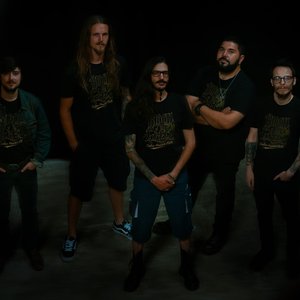 Blood of Green. Groupe de cinq hommes posant ensemble, vêtus de t-shirts noirs avec un motif doré. Leurs expressions sont sérieuses. L'homme au centre se tient avec les bras croisés, tandis que les autres entourent le groupe.