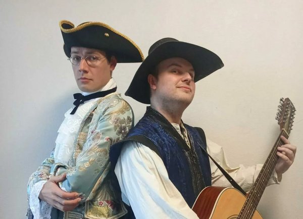 Sacré Bardel. Deux hommes posent dos à dos. L'un est vêtu d'un costume élégant avec un grand chapeau sombre, tandis que l'autre porte un gilet bleu foncé et tient une guitare. L'arrière-plan est neutre.