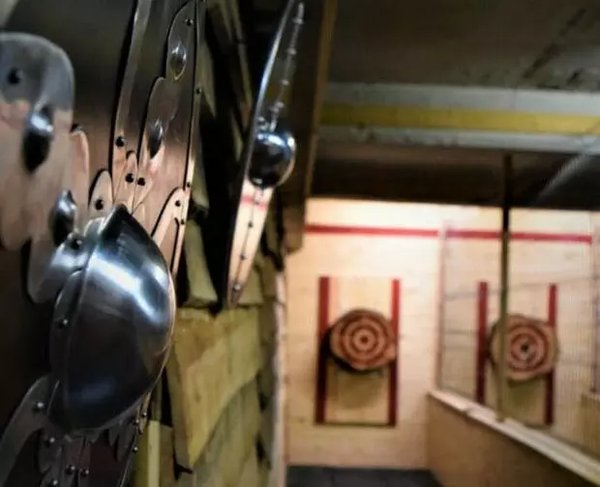 Un mur d'entraînement avec des cibles en bois, où des haches sont lancées. À gauche, on voit des protections métalliques brillantes et arrondies fixées au mur. Les cibles sont alignées à l'arrière-plan, chacune marquée d'un cercle concentrique.