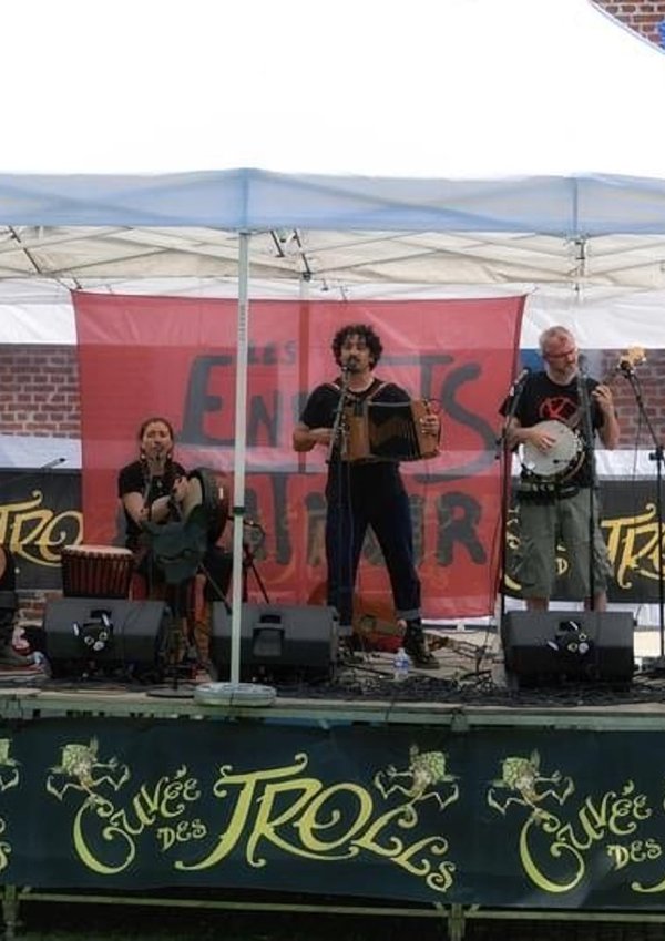 Quatre musiciens se produisent sur la scène des Estivales de Trolls et Légendes. L'un d'eux joue du violoncelle, tandis que les autres jouent de l'accordéon, du banjo et d'un instrument à vent.