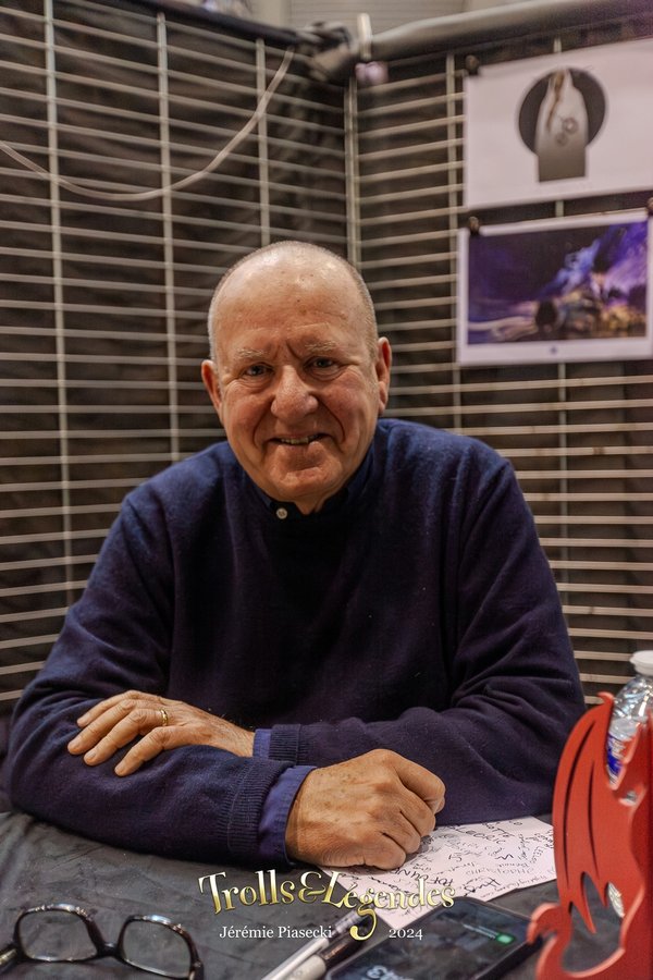 Sir Ian Livingstone, souriant, porte un pull bleu et est assis derrière une table avec des illustrations et une bouteille d'eau. Il se trouve à Trolls & Légendes en 2024