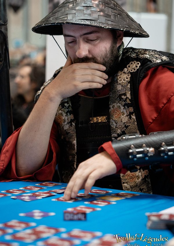 Un homme vêtu de costume ornementé et d’un chapeau conique asiatique est assis à une table de jeux. Il semble concentré, en pleine réflexion, proche des cartes et jetons sur un tapis bleu. Ambiance festival 2026.