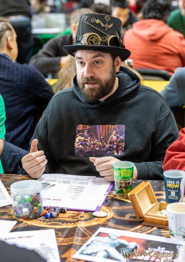 Dans le hub Jeux au Festival 2026, un homme barbu porte un chapeau orné et une capuche sombre, concentré sur une partie de société. Autour, jetons colorés, cartes et gobelets, ambiance studieuse et conviviale.