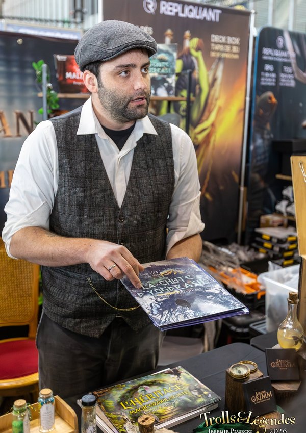 Hub Jeux au Festival 2026: un homme en gilet et casquette présente des affiches/flyers sur un stand de jeux. Derrière, des posters et produits liés à l’univers fantasy, ambiance marchande et animée.