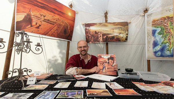 Un artiste souriant est assis à une table recouverte d'un tissu noir avec des motifs. Sur la table, il tient une bande dessinée dans une main et il y a de nombreuses illustrations étalées devant lui. À l'arrière-plan, des œuvres d'art encadrées.