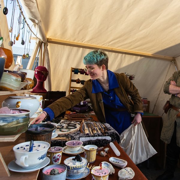 Une femme aux cheveux bleu clair, portant une veste marron et un chemisier bleu, touche des objets exposés sur une table dans un marché artisanal. Des tasses et des assiettes décoratives sont visibles, ainsi que des bijoux et des accessoires.