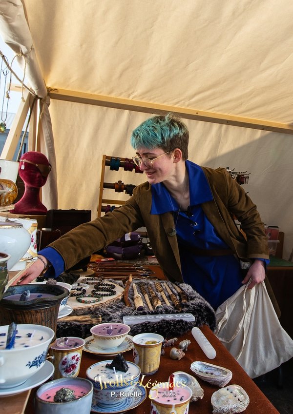 Une femme aux cheveux bleu clair, portant une veste marron et un chemisier bleu, touche des objets exposés sur une table dans un marché artisanal. Des tasses et des assiettes décoratives sont visibles, ainsi que des bijoux et des accessoires.