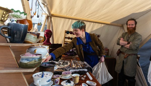 Une femme aux cheveux bleu clair, portant une veste marron et un chemisier bleu, touche des objets exposés sur une table dans un marché artisanal. Des tasses et des assiettes décoratives sont visibles, ainsi que des bijoux et des accessoires.