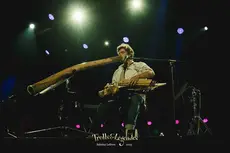 Eolya joue sur scène, un didgeridoo à la bouche et un instrument à cordes à la main. Éclairé par des lumières colorées, il est assis sur une chaise avec un micro, créant une ambiance musicale captivante lors de Trolls & Légendes 2025.