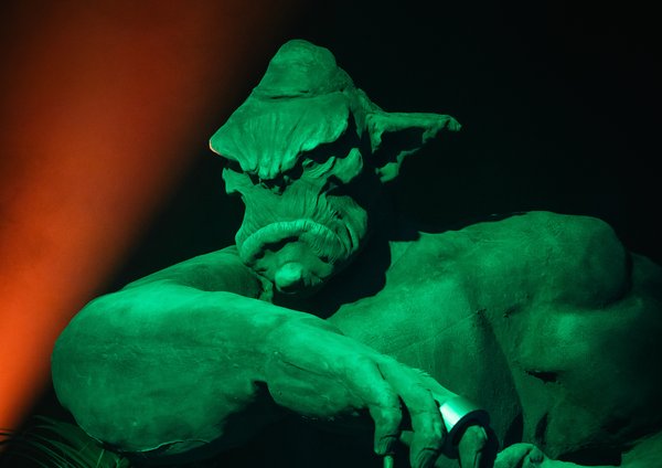 Statue d'un troll, éclairée en vert, tenant une canne, avec une expression sérieuse. L'arrière-plan est sombre, rehaussé par une lumière orange, créant une ambiance mystérieuse. Des plantes sont visibles à la base de la statue.
