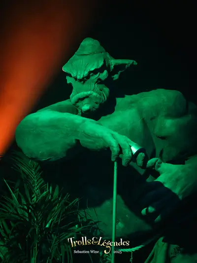 Statue d&#x27;un troll, éclairée en vert, tenant une canne, avec une expression sérieuse. L&#x27;arrière-plan est sombre, rehaussé par une lumière orange, créant une ambiance mystérieuse. Des plantes sont visibles à la base de la statue.