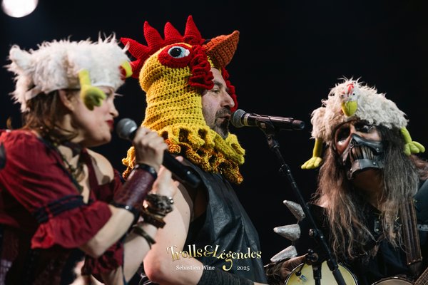 Naheulband en 2025, habillés de costumes amusants : l'un porte un chapeau en crochet en forme de poule, les autres portent des accessoires en plumes. Ils chantent dans un ambiance festive, avec des micros en main.