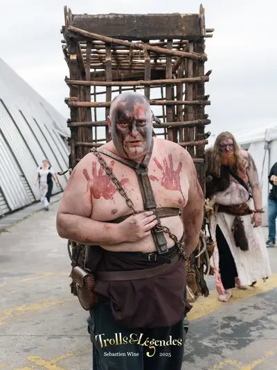 Un homme musclé, torse nu, avec des taches de peinture rouge sur le corps, porte un harnais et semble être un personnage de type médiéval. Il se tient devant une structure en bois, semblable à une cage, qui est poussée par une autre personne à l'arrière.