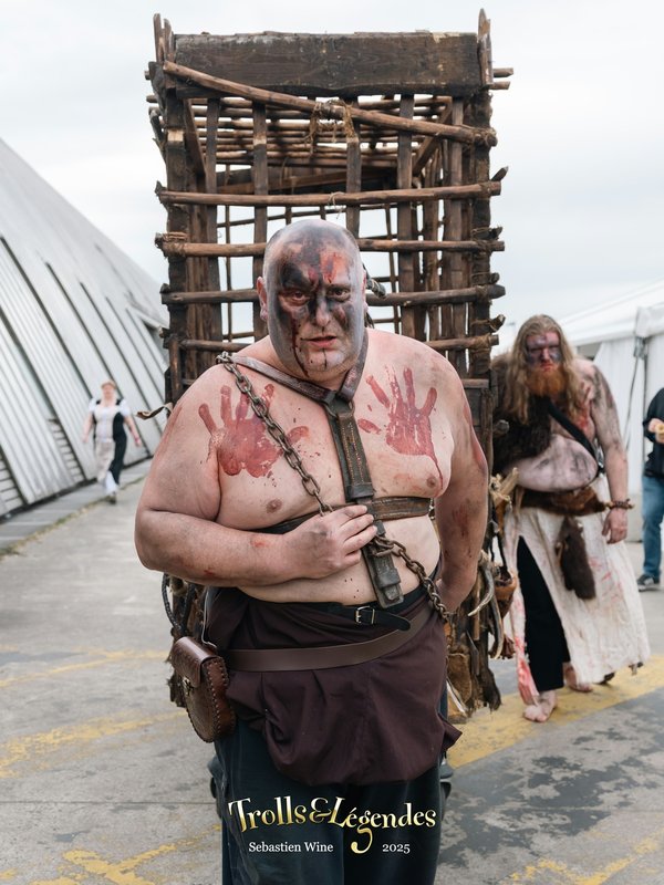 Un homme musclé, torse nu, avec des taches de peinture rouge sur le corps, porte un harnais et semble être un personnage de type médiéval. Il se tient devant une structure en bois, semblable à une cage, qui est poussée par une autre personne à l'arrière.