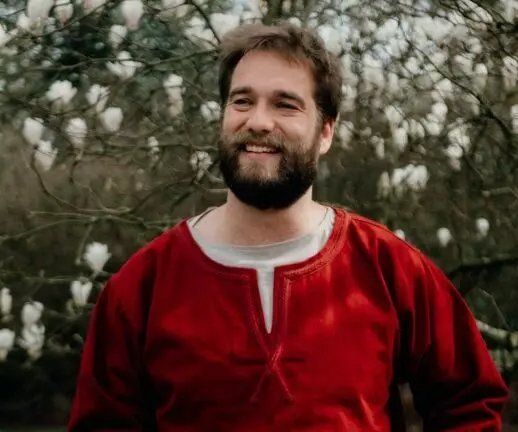 Un homme souriant porte une tunique rouge avec un col en V. Il a une barbe et des cheveux bruns, et il se tient devant un arbre avec des fleurs blanches en arrière-plan. L'image dégage une atmosphère joyeuse et naturelle.