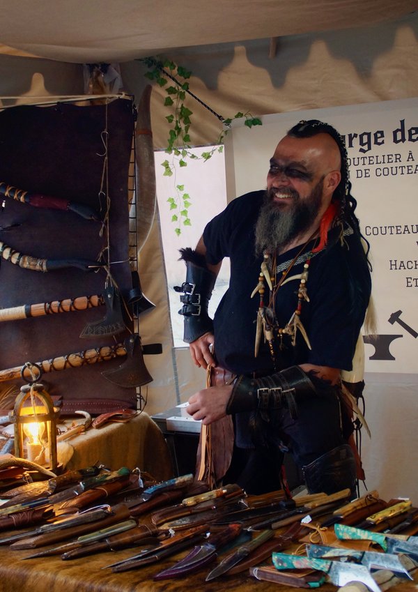 Artisan costumé souriant derrière un étal de couteaux et lanternes au stand Trolls & Légendes.