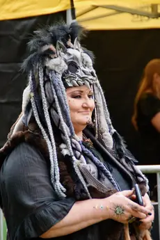 Een vrouw gekleed in een donkere outfit draagt een indrukwekkende cosplay met een pluchen wolfshoofd versierd met vele vlechten. Ze staat voor een gele tent, met een vredige uitstraling. Het evenement vindt plaats tijdens de Estival...