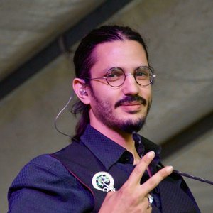 Op het podium van de Estivales 2025 komt de groep 'Blood of Green' energiek naar voren. De zanger, met ronde brillen en een donker shirt, toont een zelfverzekerde glimlach terwijl hij een vredesteken maakt. De muzikale sfeer is veelbelovend.