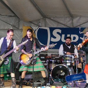 Op het podium treedt de band 'Blood of Green' op tijdens de Estivales 2025. De muzikanten, gekleed in kilts, spelen vol energie met elektrische gitaar, bas en viool. De feestelijke sfeer wordt versterkt door de simpele decoratie en de versterkers op de...