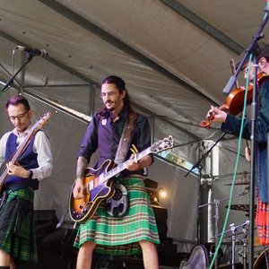 Op het podium tijdens de Estivales 2025, treedt de band "Blood of Green" energiek op. De muzikanten dragen Schotse kilts en spelen gitaar, viool en een ander instrument. De sfeer is feestelijk onder een tent en boeit het publiek.