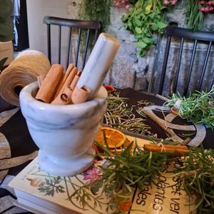 Mortier blanc avec bâtons de cannelle, herbes séchées et fleurs, ambiance atelier magique.