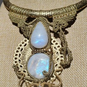 Les 1000 et un nœuds. Collier en macramé ornée de pierres précieuses en forme de goutte, dont deux en opale brillante, avec un détail en métal doré en arrière-plan. Le col est tressé et l'ensemble dégage une élégance artisanale.