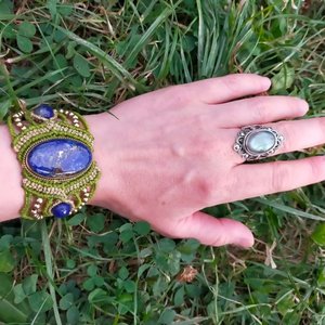 Les 1000 et un noeuds. Une main portant un bracelet en macramé vert avec une pierre ovale bleue au centre, entourée de petites perles. À côté, un anneau en argent avec une pierre ovale de couleur pâle. L'image est prise sur un fond de gazon vert.