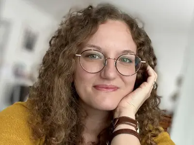 Célia BEAUDUC - Portrait d'une femme souriante avec des lunettes et des cheveux bouclés.