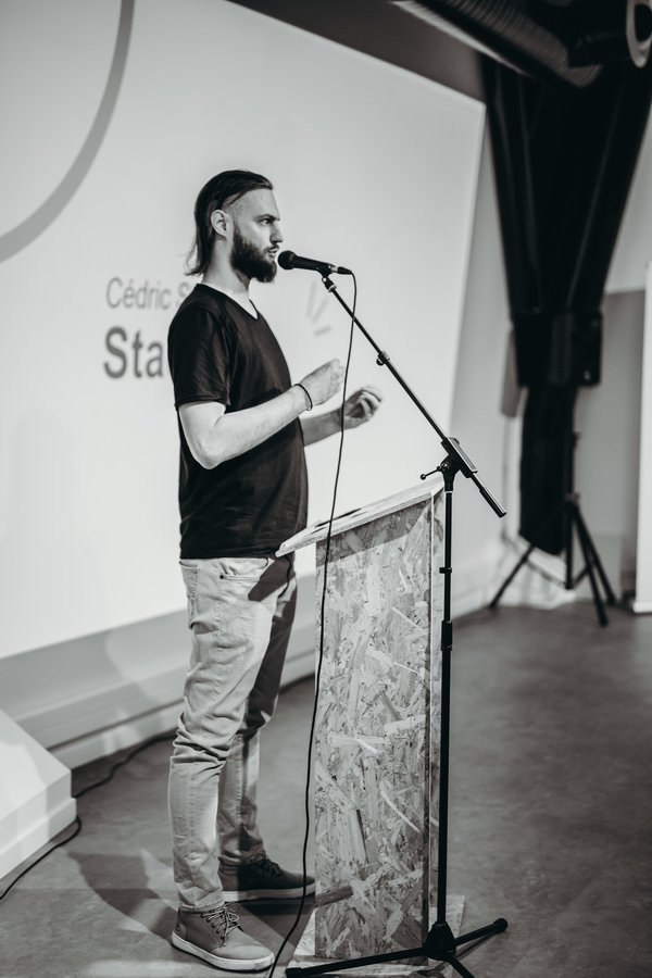 Cédric Staces. Un homme debout sur une scène, parlant au microphone.